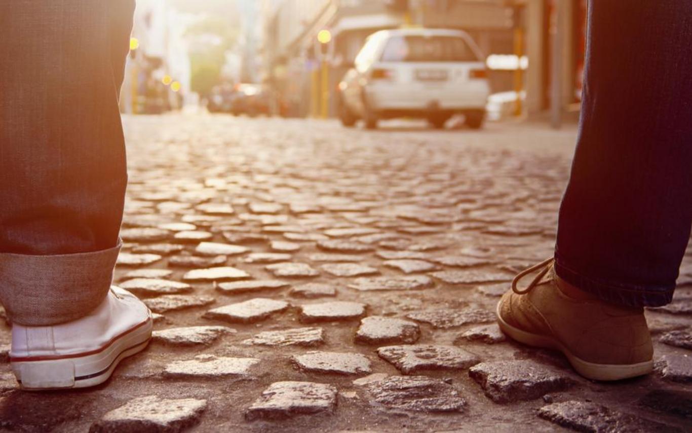 Close-up of two people walking side by side on a cobblestone street at sunset, with warm light reflecting off the stones and city buildings in the background.